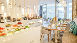 A modern dining area with a buffet, long tables, and a seating area with cushioned chairs. - Grand Mercure Dubai City (Dubai)