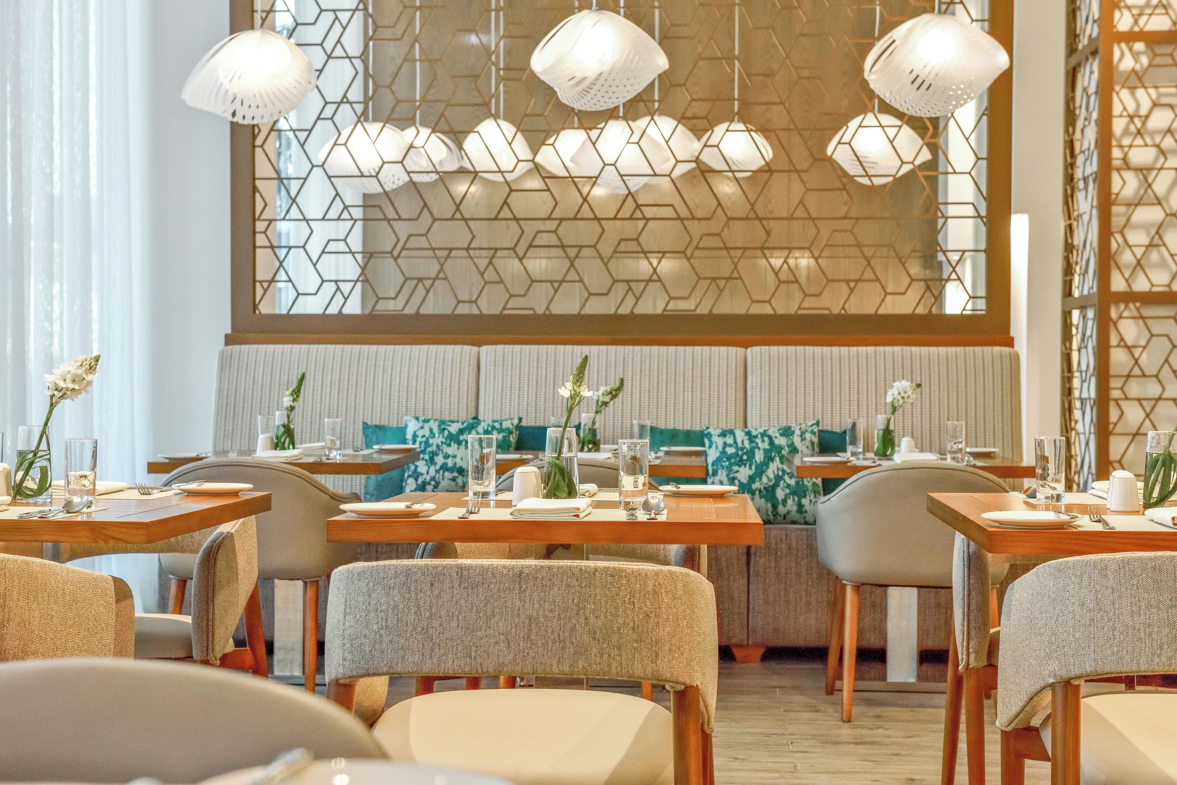A restaurant with wooden tables and chairs, hexagonal patterned wall, hanging pendant lights, and green and white cushions on the seating.