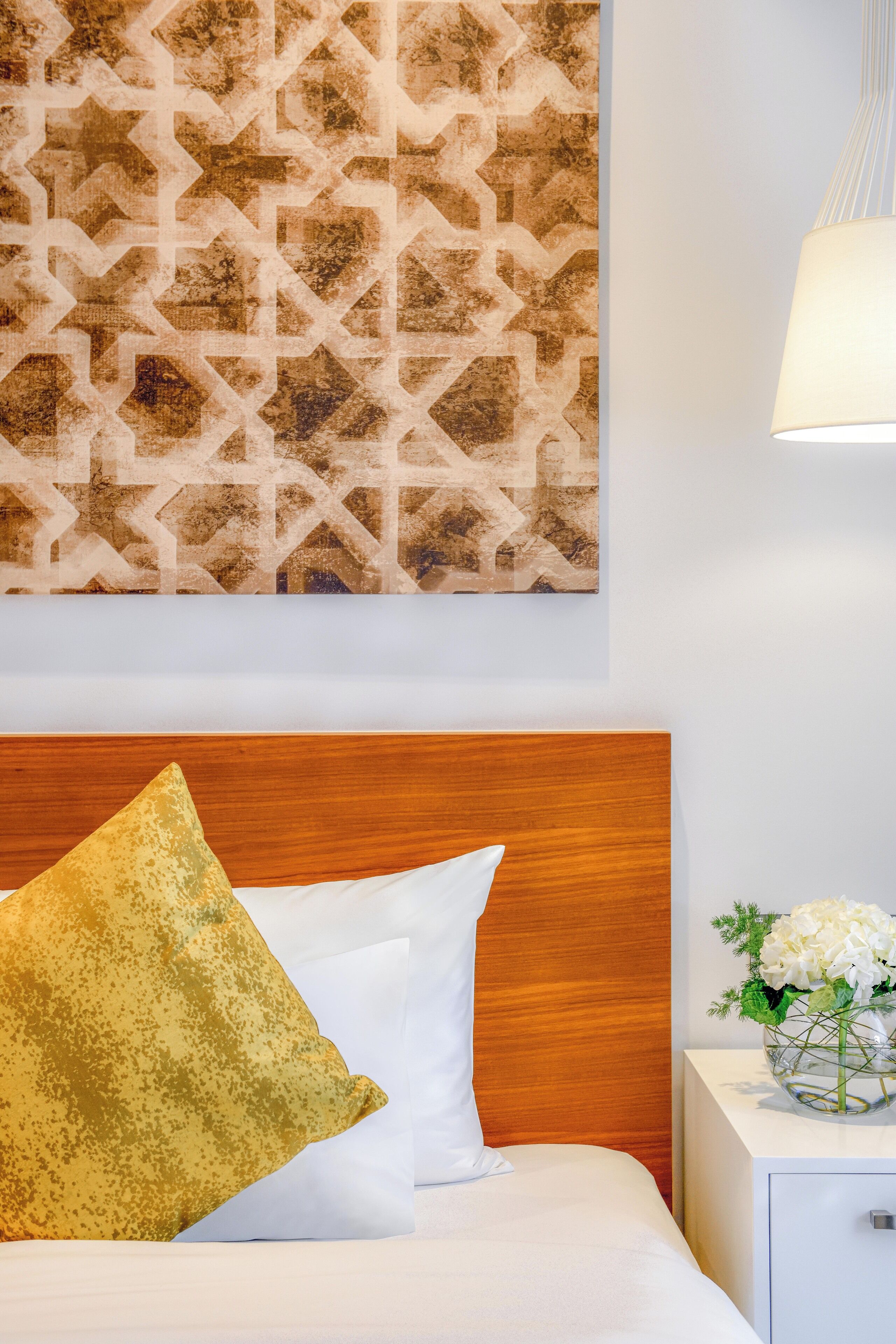 A neatly made bed with a wooden headboard, a decorative wall piece, and a bedside table with a vase of flowers.
