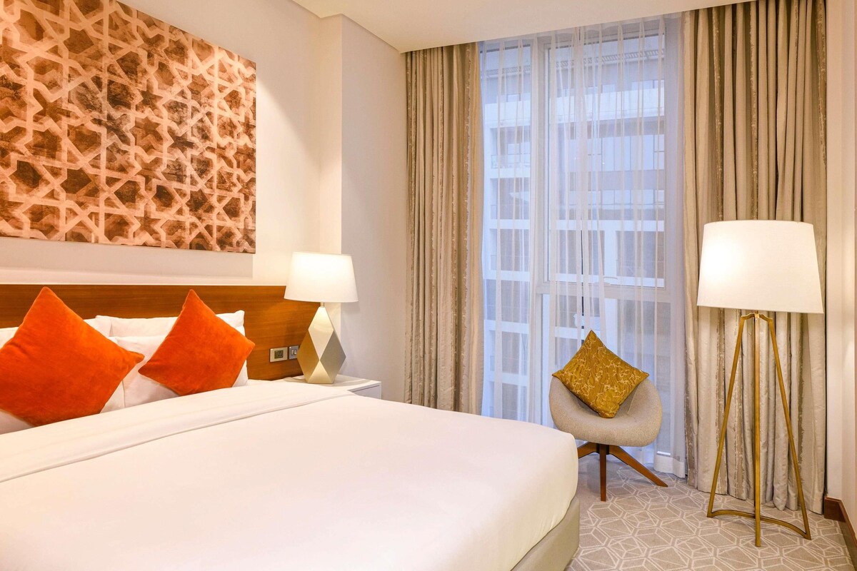 A hotel room with a large bed, two bedside lamps, a chair, a large window with curtains, and a geometric-patterned wall.