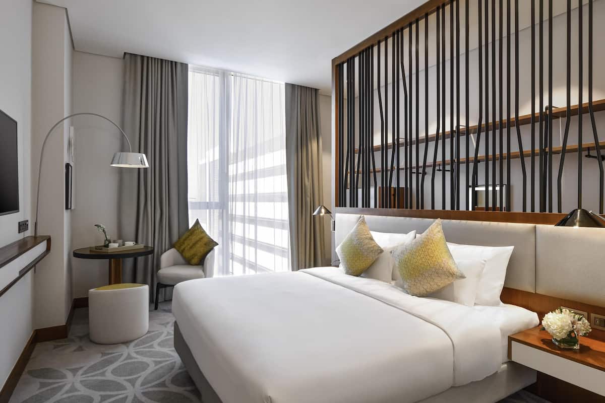 Une chambre d’hôtel moderne, dotée d’un grand lit, d’un bureau, d’une chaise, d’un lampadaire et d’une grande fenêtre avec des rideaux.