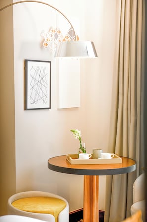 A hotel room with a round table, a framed abstract artwork, a lamp, and a window with curtains.
