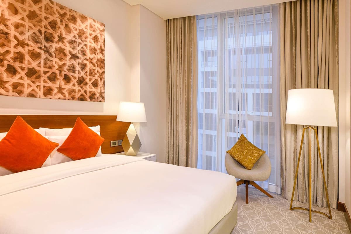 Une chambre d’hôtel avec un grand lit, deux lampes de chevet, une chaise, une grande fenêtre avec des rideaux et un mur orné d’un motif géométrique.