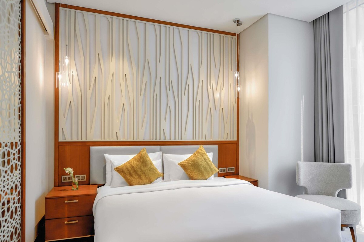 A hotel room with a large bed, a wooden headboard, a nightstand, a chair, and a decorative wall panel.