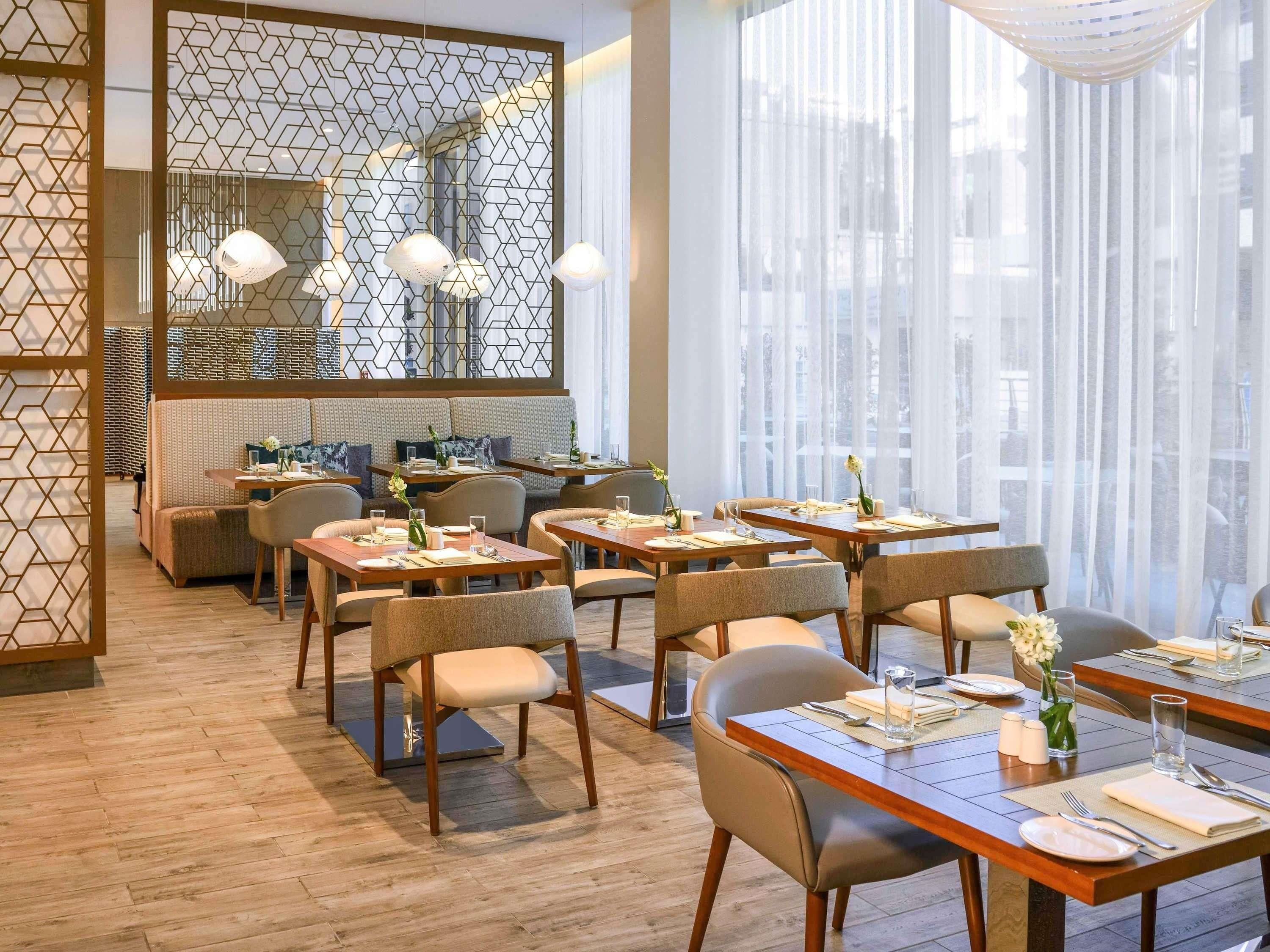 A modern restaurant interior with wooden flooring, round tables, cushioned seating, and a large mirror.