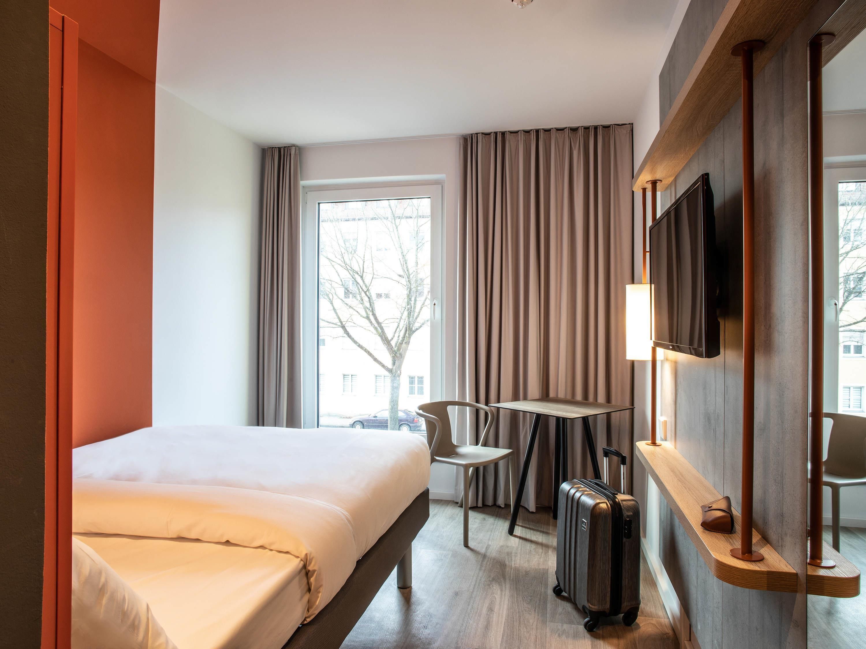 Habitación de hotel con cama, escritorio, silla, televisión y maleta.