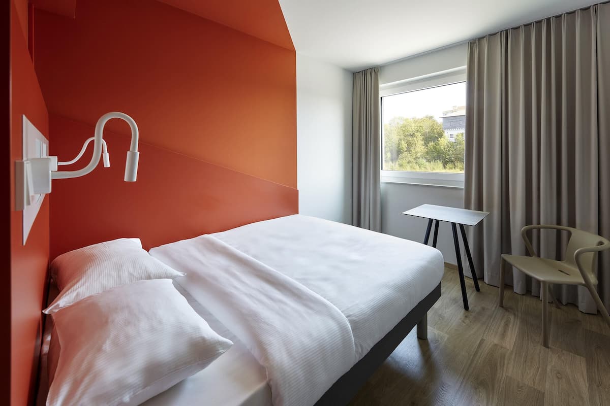 A hotel room with a bed, a small table, a chair, and a window with curtains.