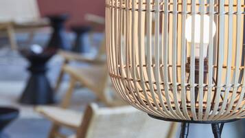 A wicker lamp with a visible light bulb inside, placed on a table with a blurred background of chairs and tables.