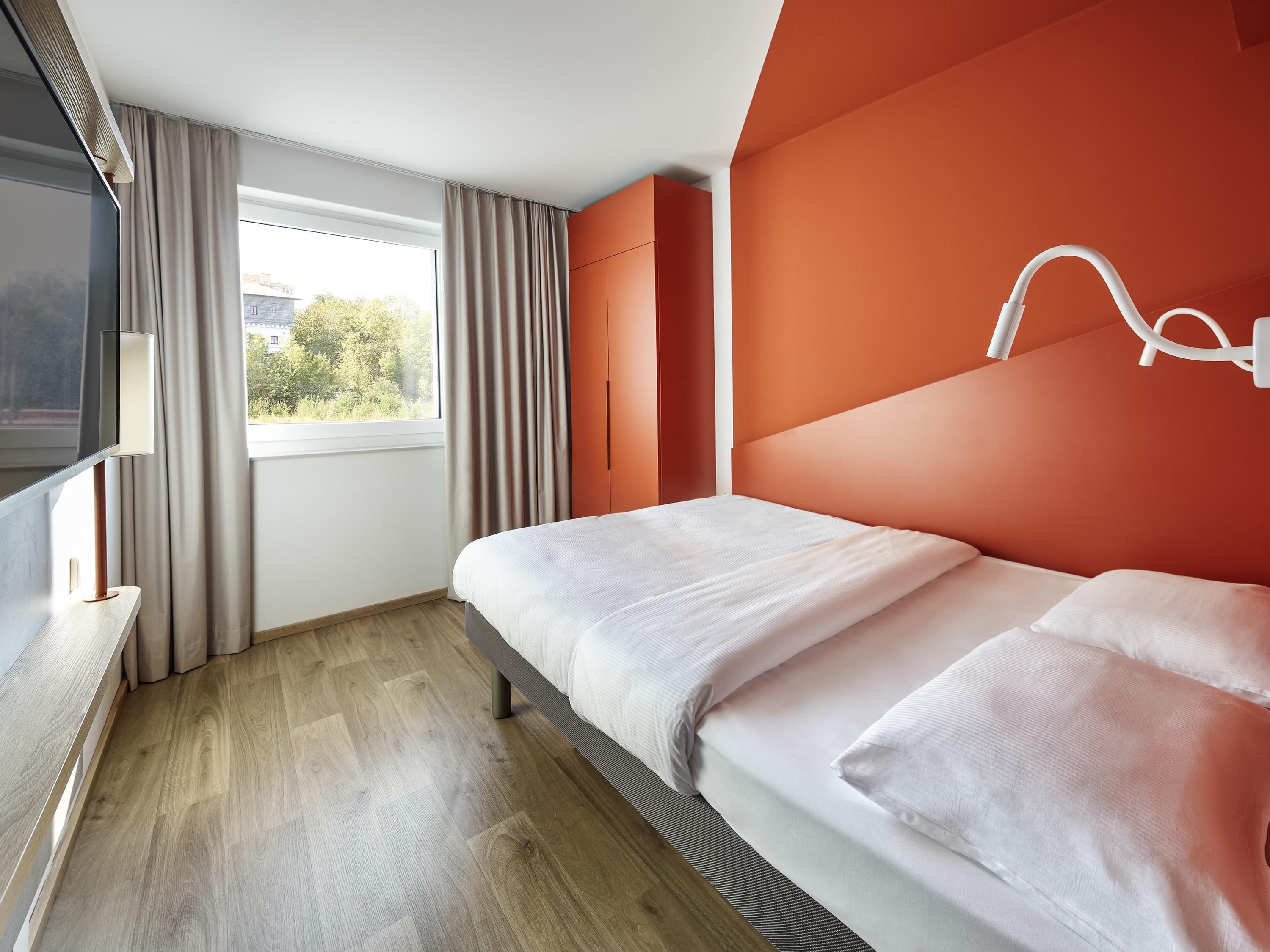 A modern hotel room with a bed, a TV, a window with curtains, and a view of greenery outside.