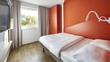 A modern hotel room with a bed, a TV, a window with curtains, and a view of greenery outside.
