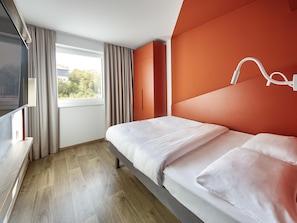 A modern hotel room with a bed, a TV, a window with curtains, and a view of greenery outside.