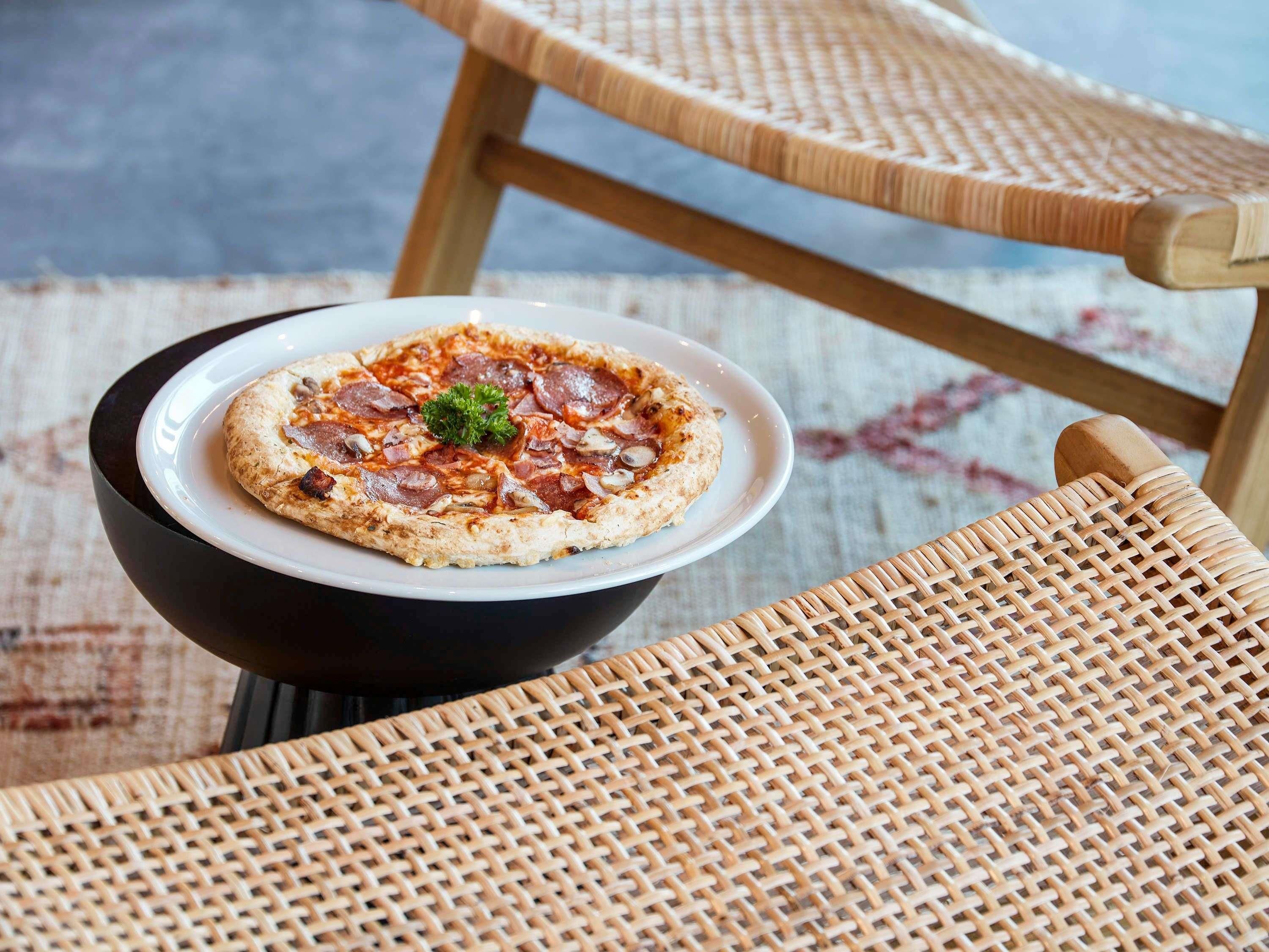 A pizza on a white plate on a black bowl, placed on a wicker chair.
