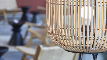 A wicker lamp with a visible light bulb inside, placed on a table with a blurred background of chairs and tables.