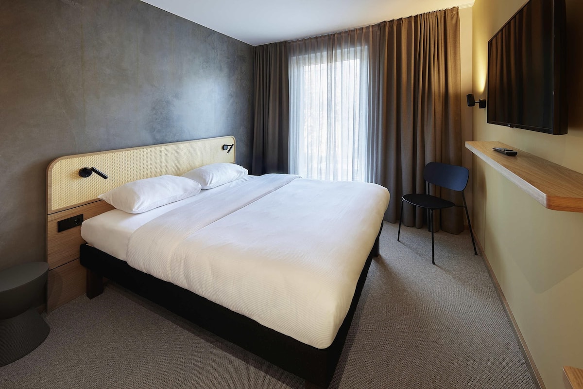 A modern hotel room with a large bed, a desk with a chair, a television, and a window with curtains.