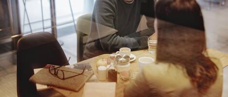 Una persona sentada a una mesa con una taza y un vaso de agua, en una habitación con una silla y una mesa.