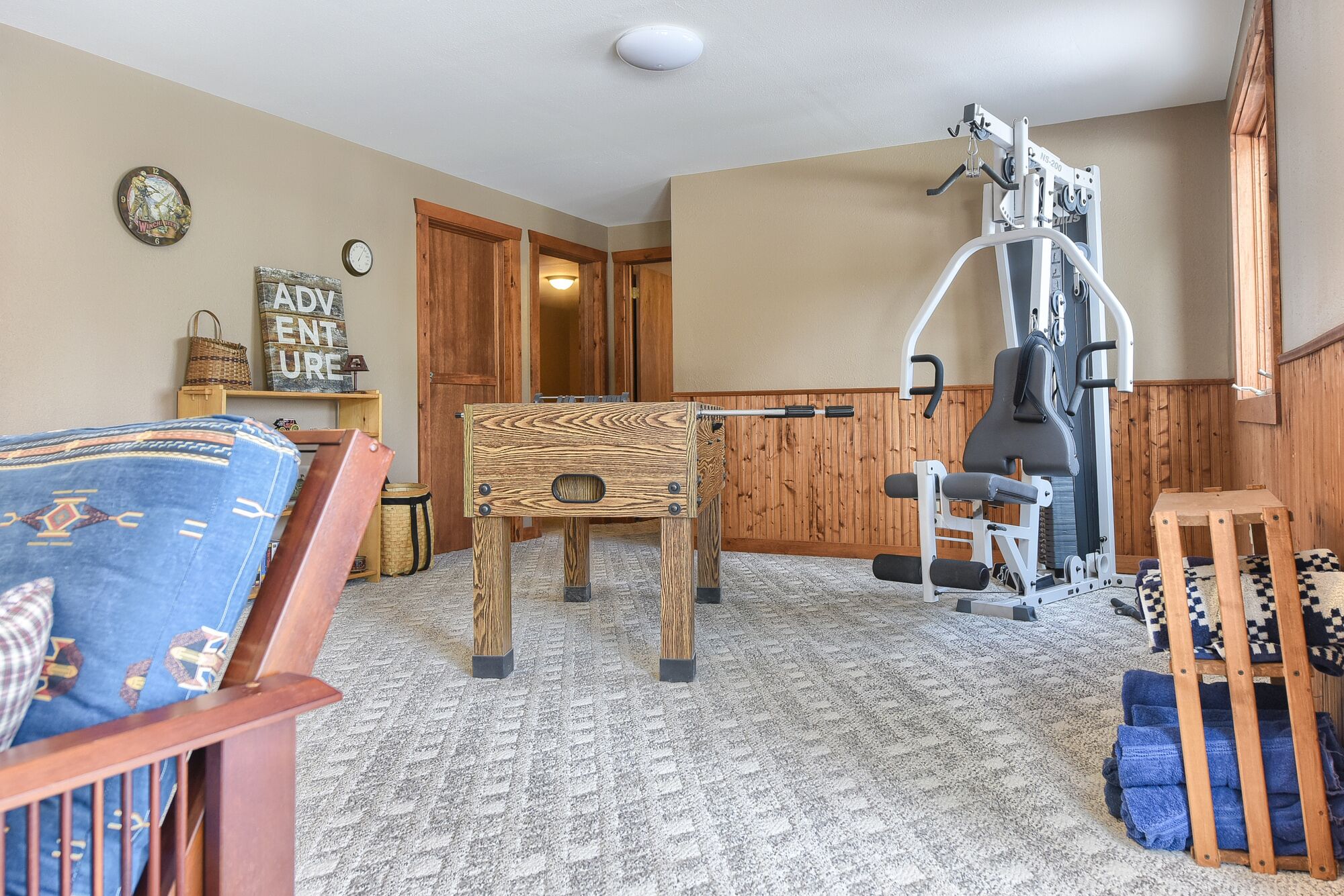 Ein Zimmer mit einem Holztisch, Trainingsgeräten, einem blau gemusterten Sofa und einer Wanduhr.