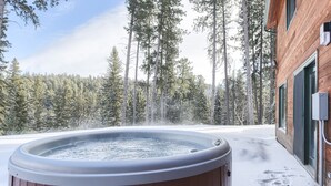 Ett bubbelbad utomhus, omgivet av snö, med en skog i bakgrunden och en byggnad av trä.