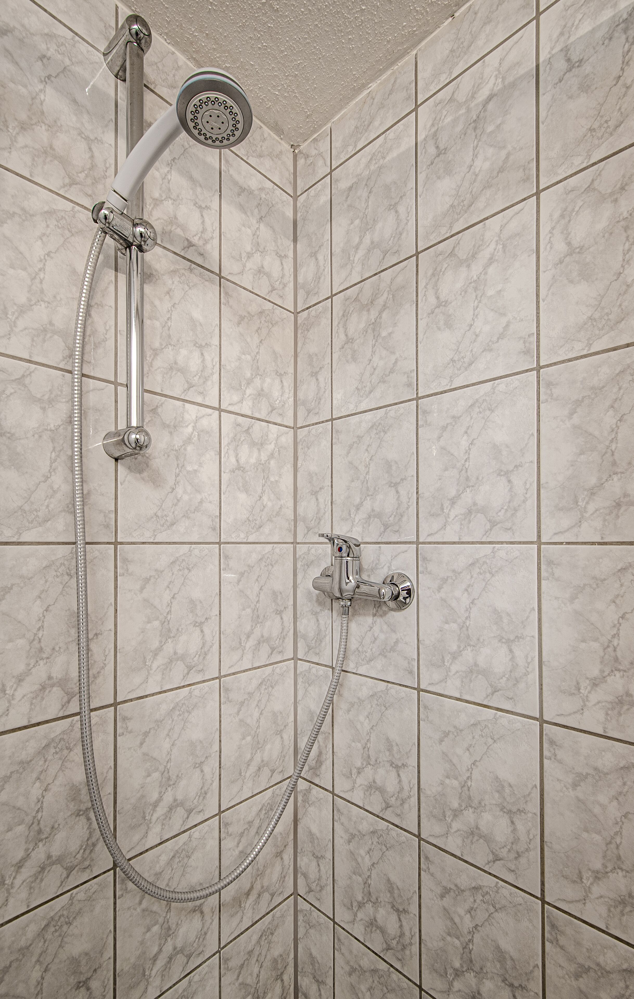 A shower with a handheld showerhead and a wall-mounted faucet, set against a tiled wall with a marble pattern.