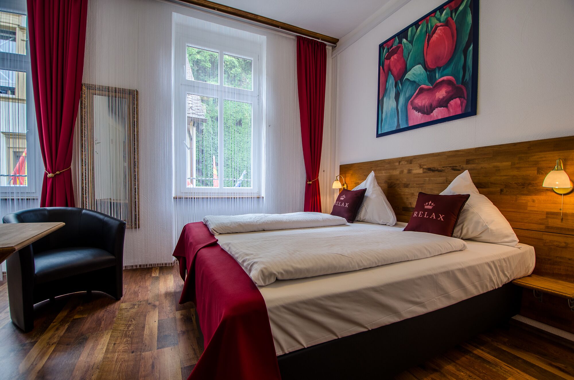 A hotel room with a double bed, two pillows with 'RELAX' text, a wooden headboard, a table, a chair, and a large window with red curtains.