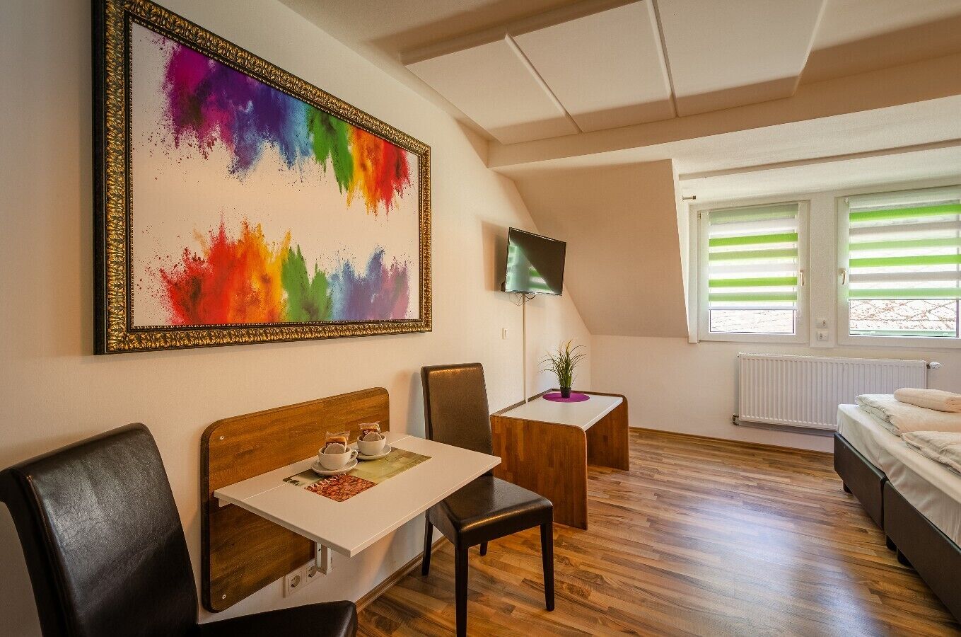 Habitación de hotel con piso de madera, una cama, un escritorio con silla, televisor y un cuadro abstracto grande en la pared.