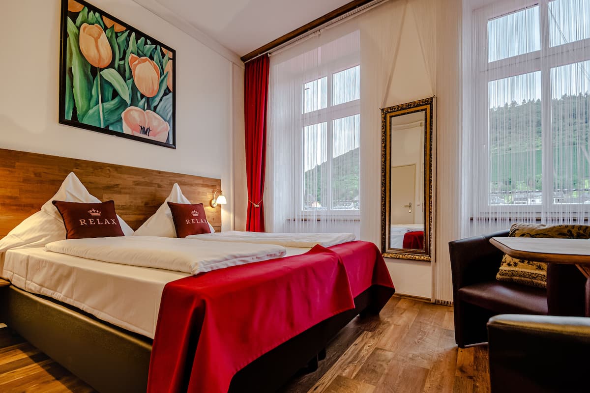Ein Hotelzimmer mit einem großen Bett, einer roten Bettdecke, einem Gemälde mit Tulpen, einem Spiegel und Blick auf Bäume durch das Fenster.