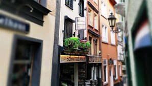 Eine schmale Straße, gesäumt von mehrstöckigen Gebäuden mit unterschiedlichen Architekturstilen und Hinweisschildern für Geschäfte.