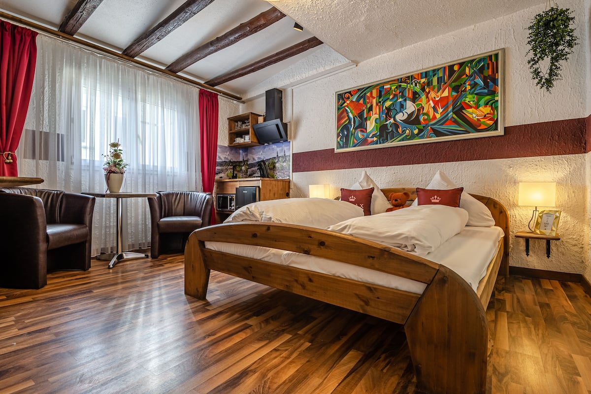 Ein hölzernes Bett mit weißer Bettwäsche, ein roter Vorhang, ein kleiner Tisch mit Pflanze und ein farbenfrohes, abstraktes Gemälde an der Wand.