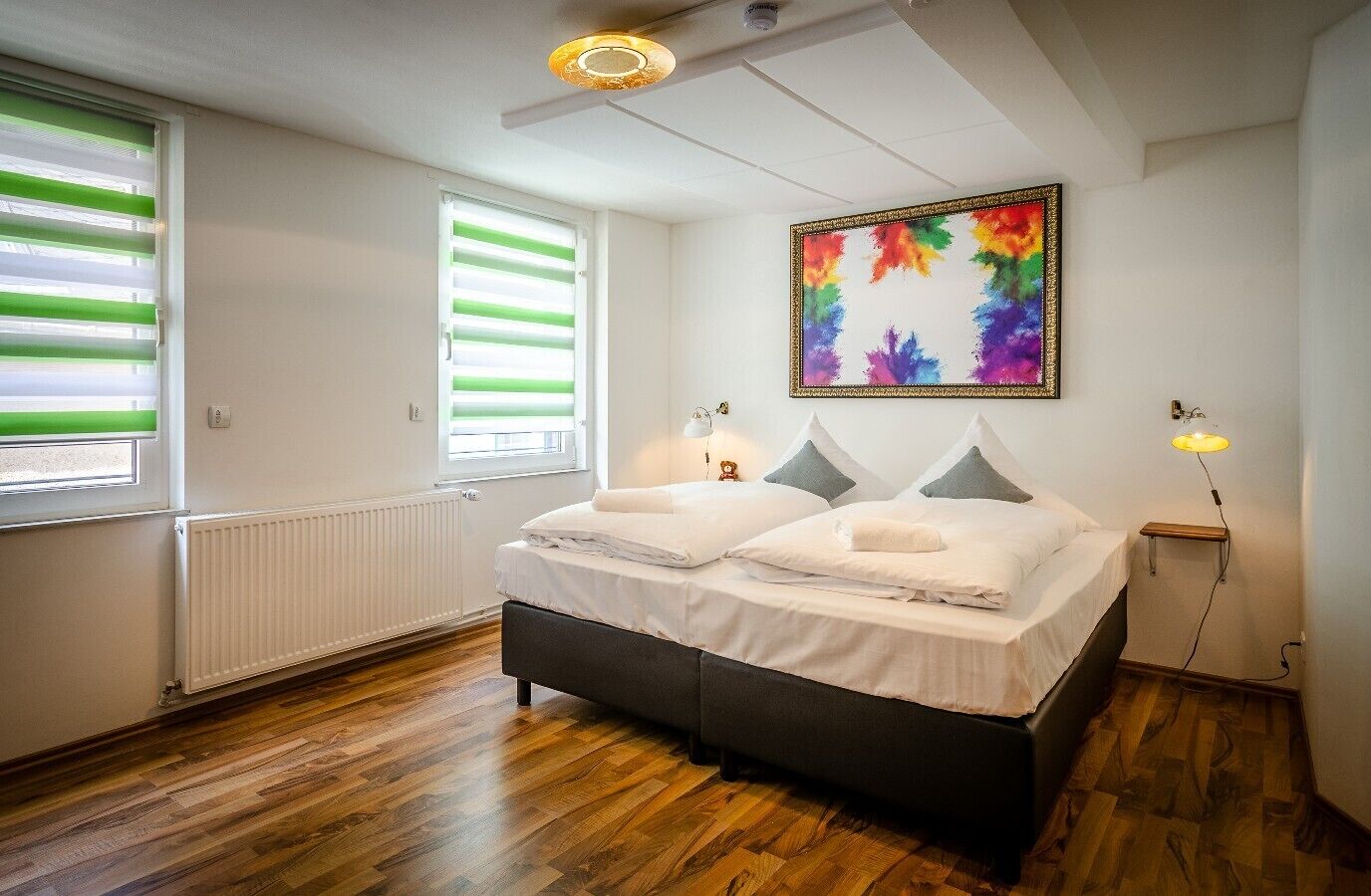 A bedroom with a bed, two bedside tables, a radiator, a window with blinds, a ceiling light, and a colorful painting on the wall.