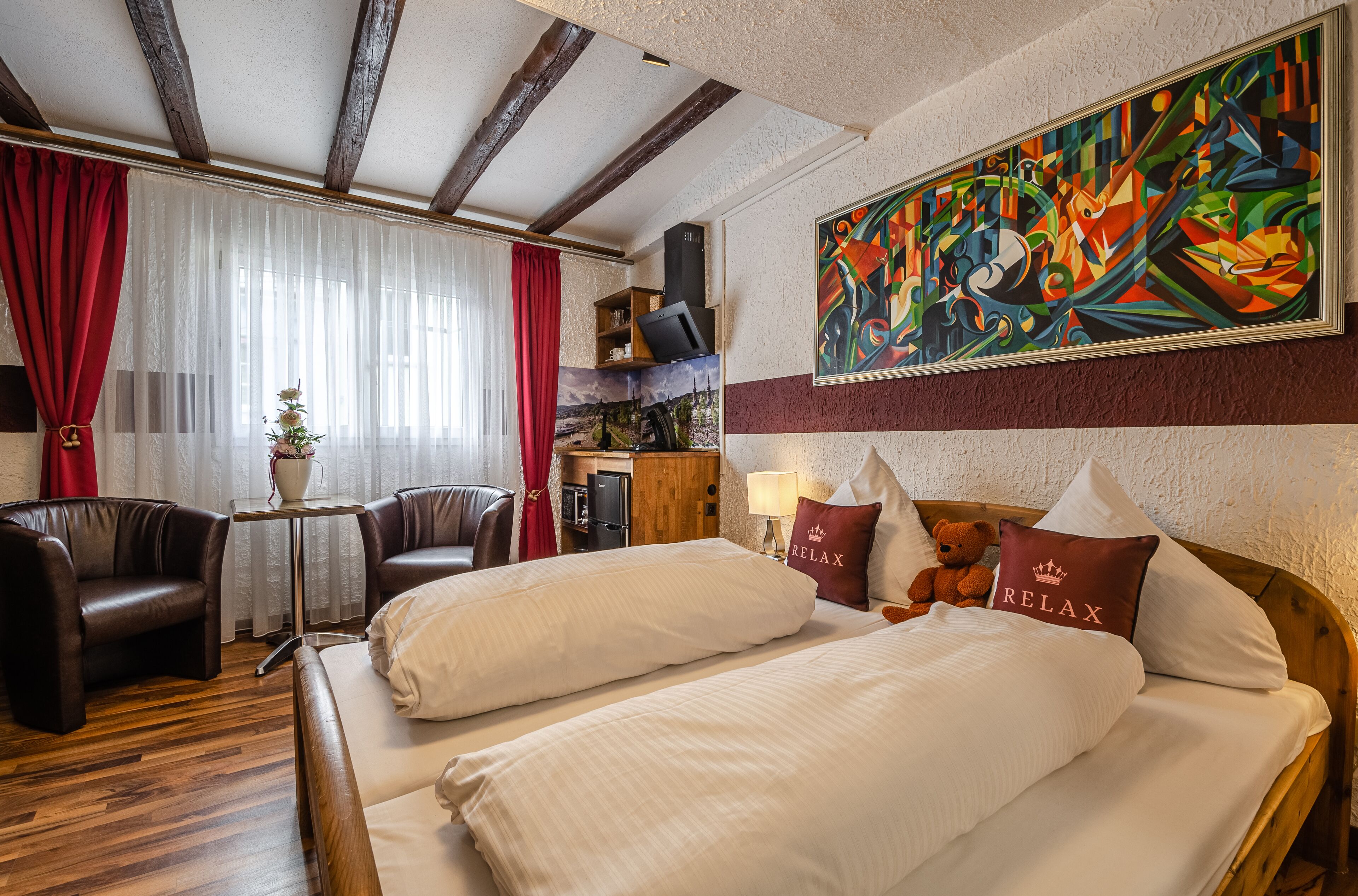 A hotel room with a wooden ceiling, a bed with white bedding, two pillows with 'RELAX' text, a teddy bear, a red curtain, a small table with a plant, and a colorful abstract painting on the wall.