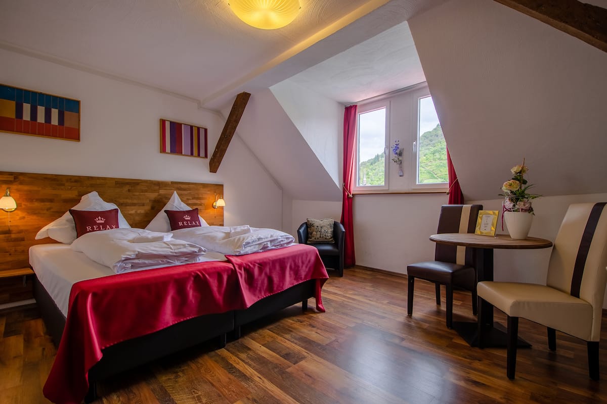 Ein Schlafzimmer mit einem hölzernen Kopfteil, einem Bett mit roter Bettwäsche, einem kleinen Tisch mit Stühlen und einem Fenster mit Blick auf Grünflächen.