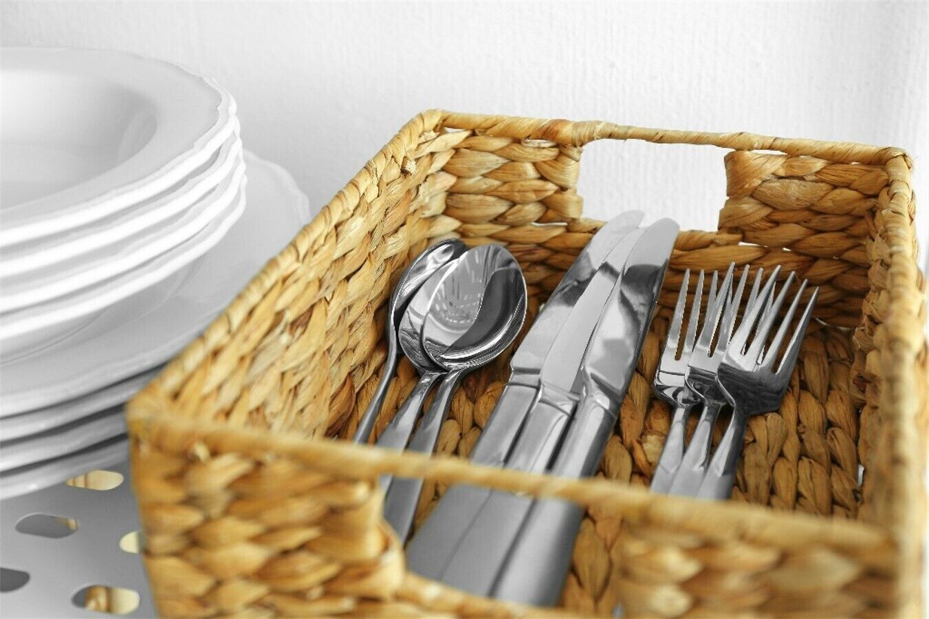 A woven basket containing a spoon, knife, and fork, placed on a surface with stacked white plates in the background.