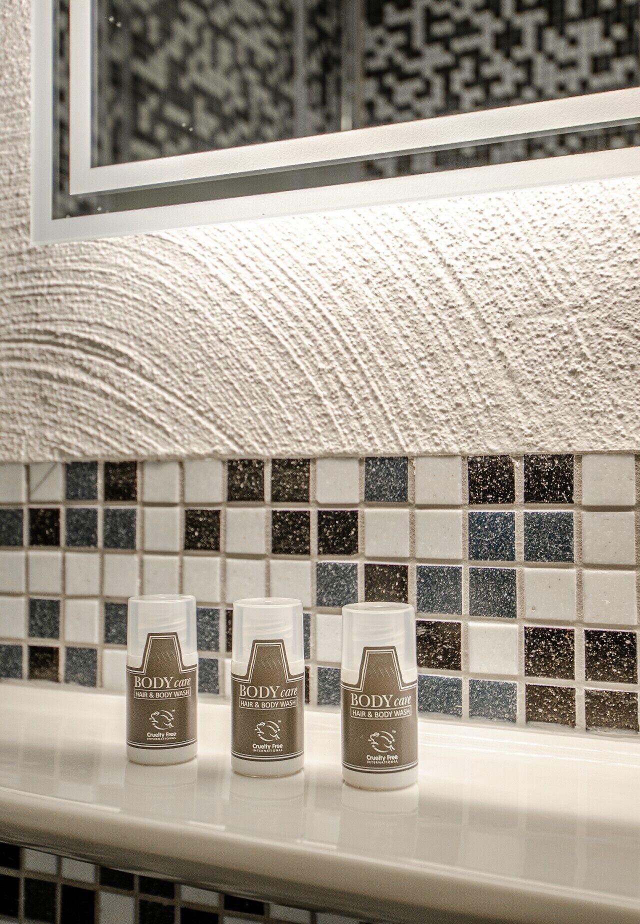 Three bottles of Body Care hair and body wash on a bathroom counter with a tiled backsplash.