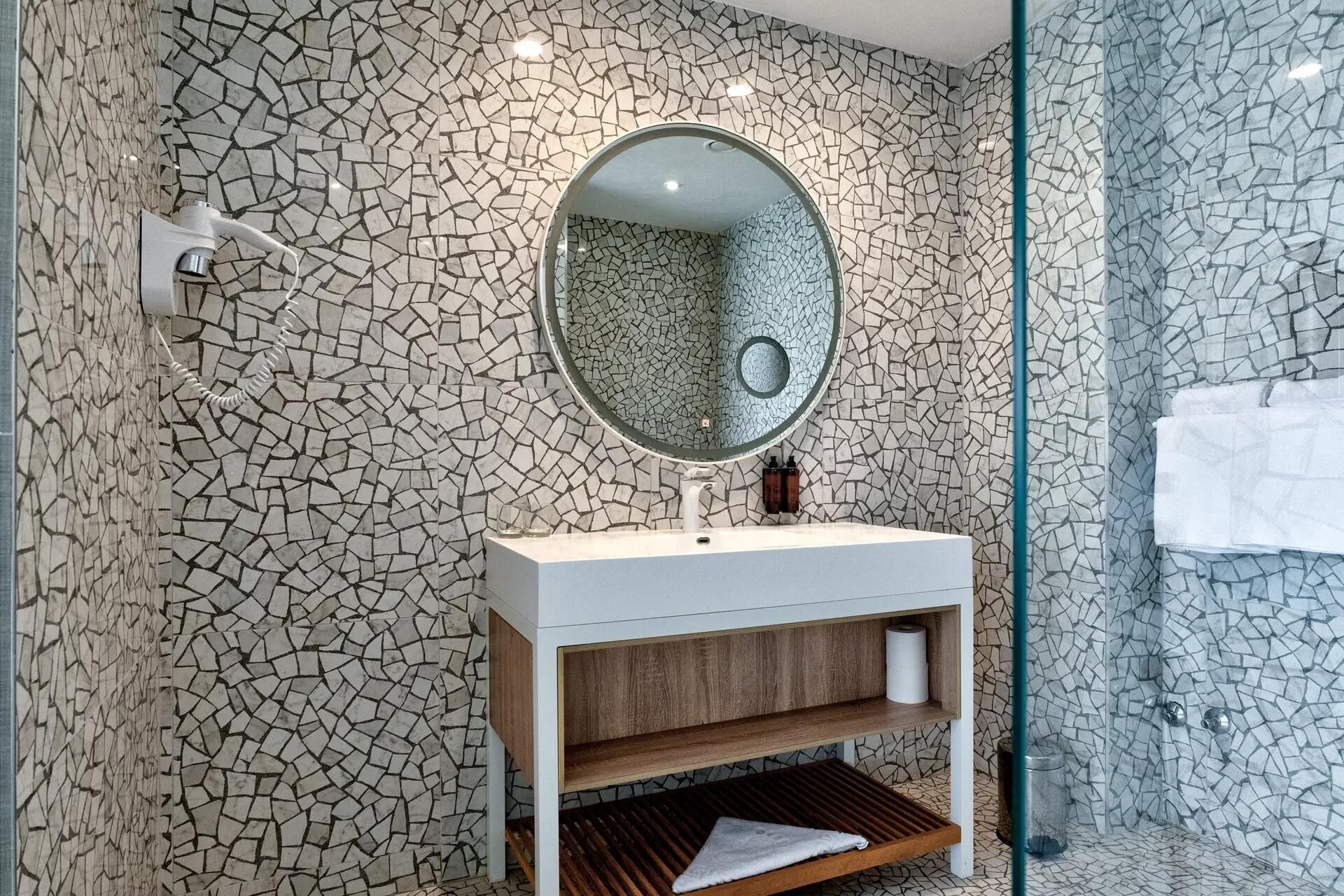A bathroom with a mosaic tile wall, a round mirror, a white sink, and a shower area.