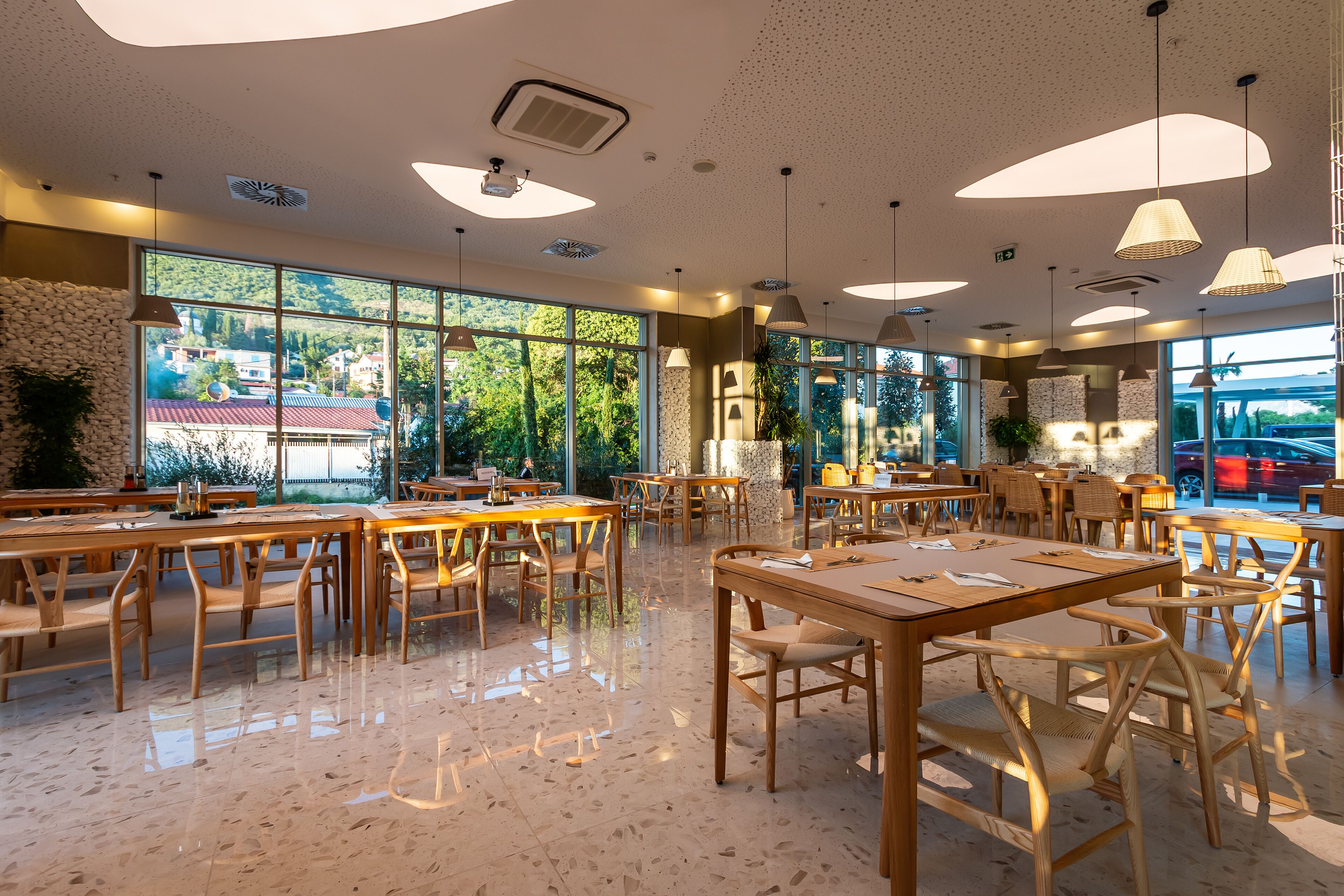 A spacious restaurant with wooden tables and chairs, large windows offering a view of the outdoors, and modern pendant lighting.