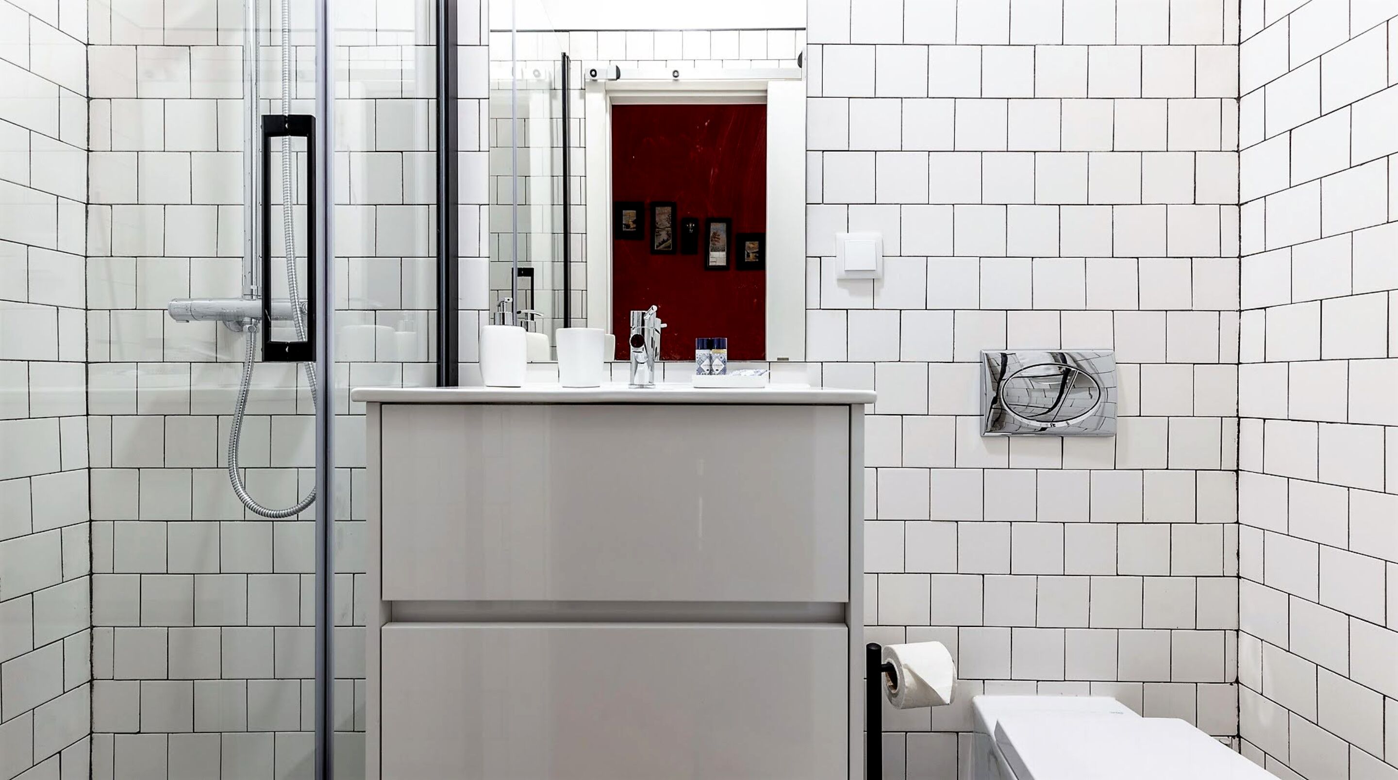 A modern bathroom with white tiled walls, a glass shower enclosure, a white sink with a mirror, and a red accent wall.