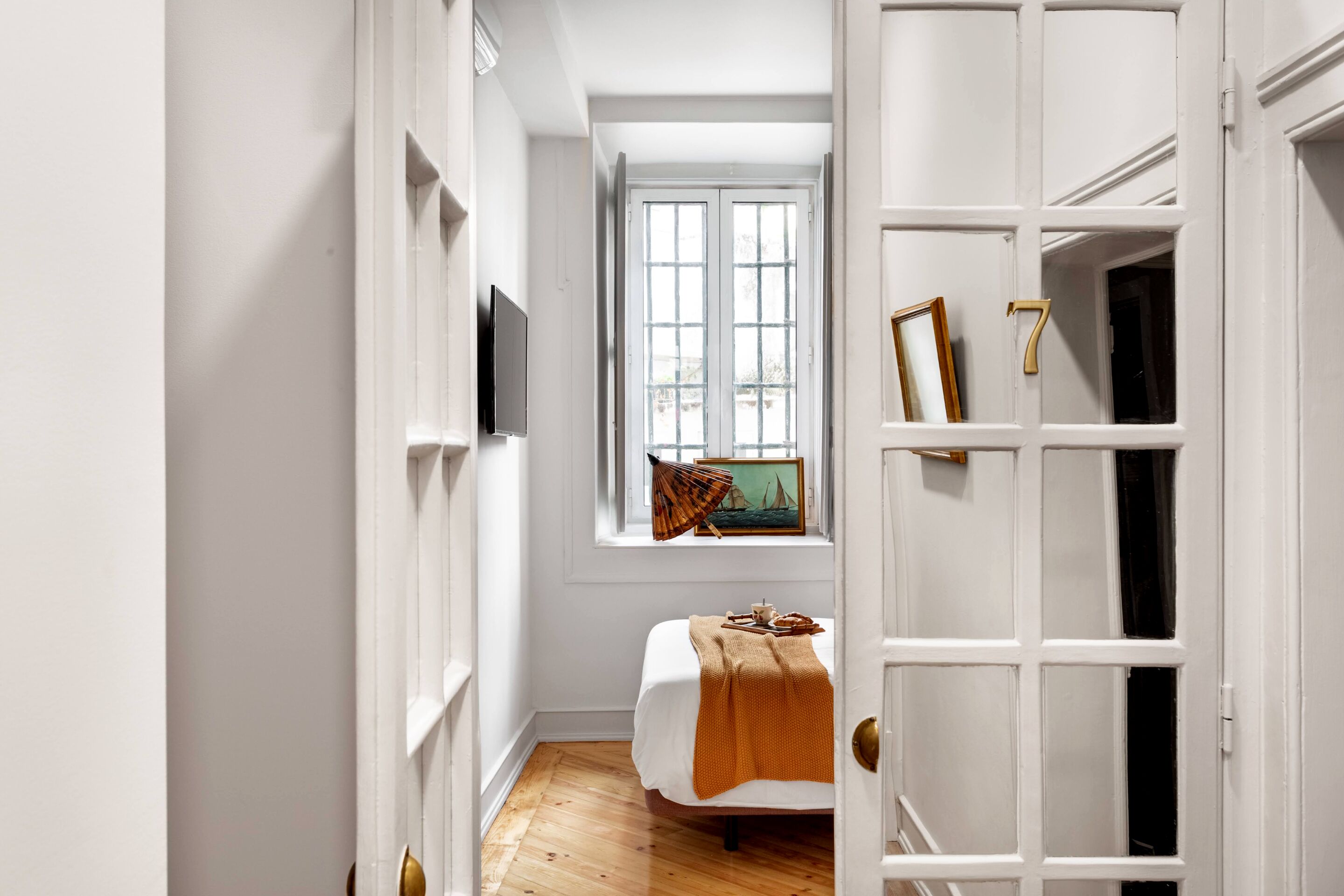 Un pasillo estrecho con una cama al final, una ventana con vistas y una puerta con un número.