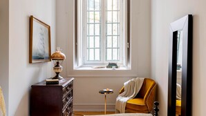 A room with a large window, a wooden cabinet, a chair, and a small table with books and a lamp. - LeL Madalena GuestHouse (Lisbon)