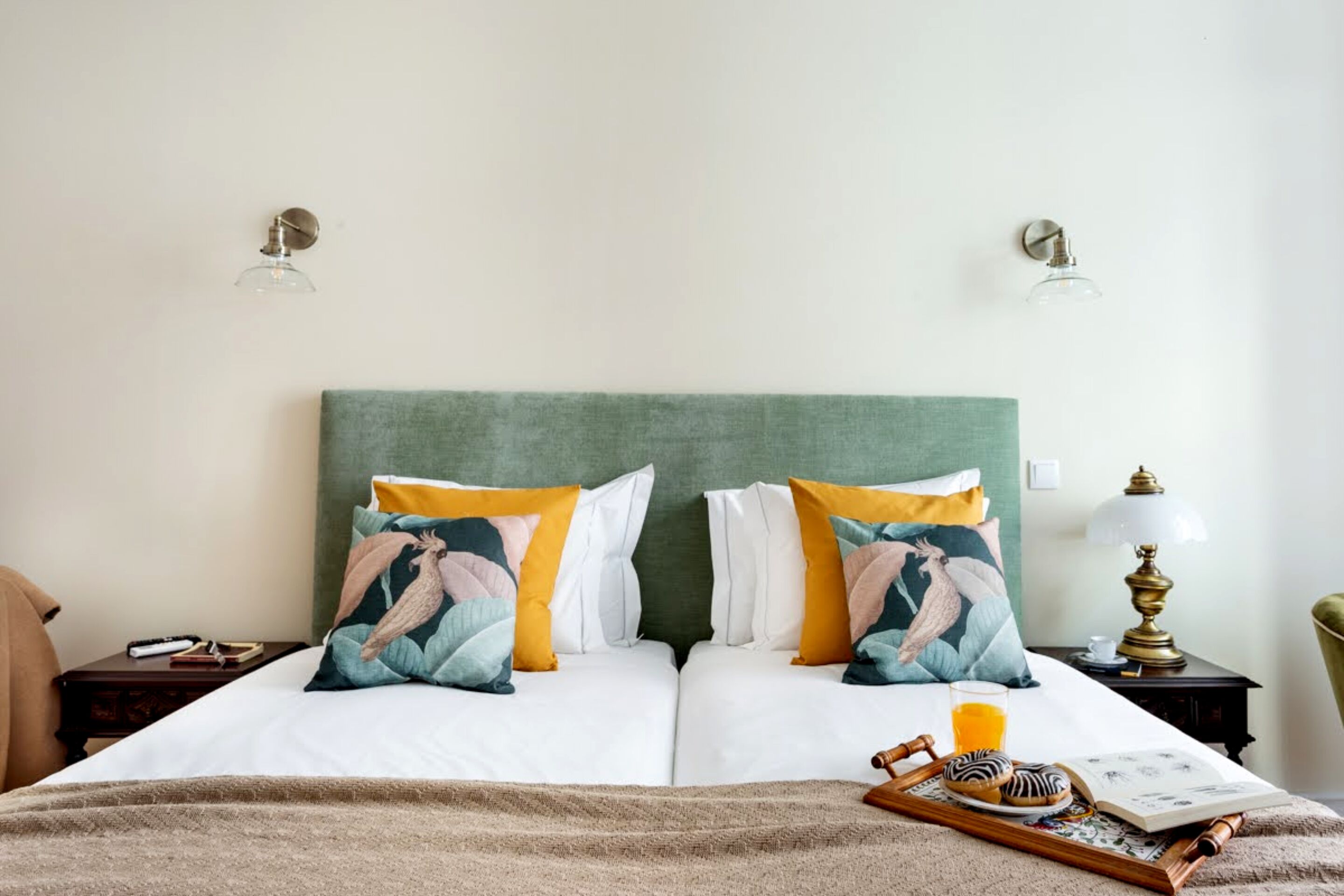 A neatly made double bed with two pillows, a bedside table with a lamp, a tray with cookies and a book, and a glass of orange juice.