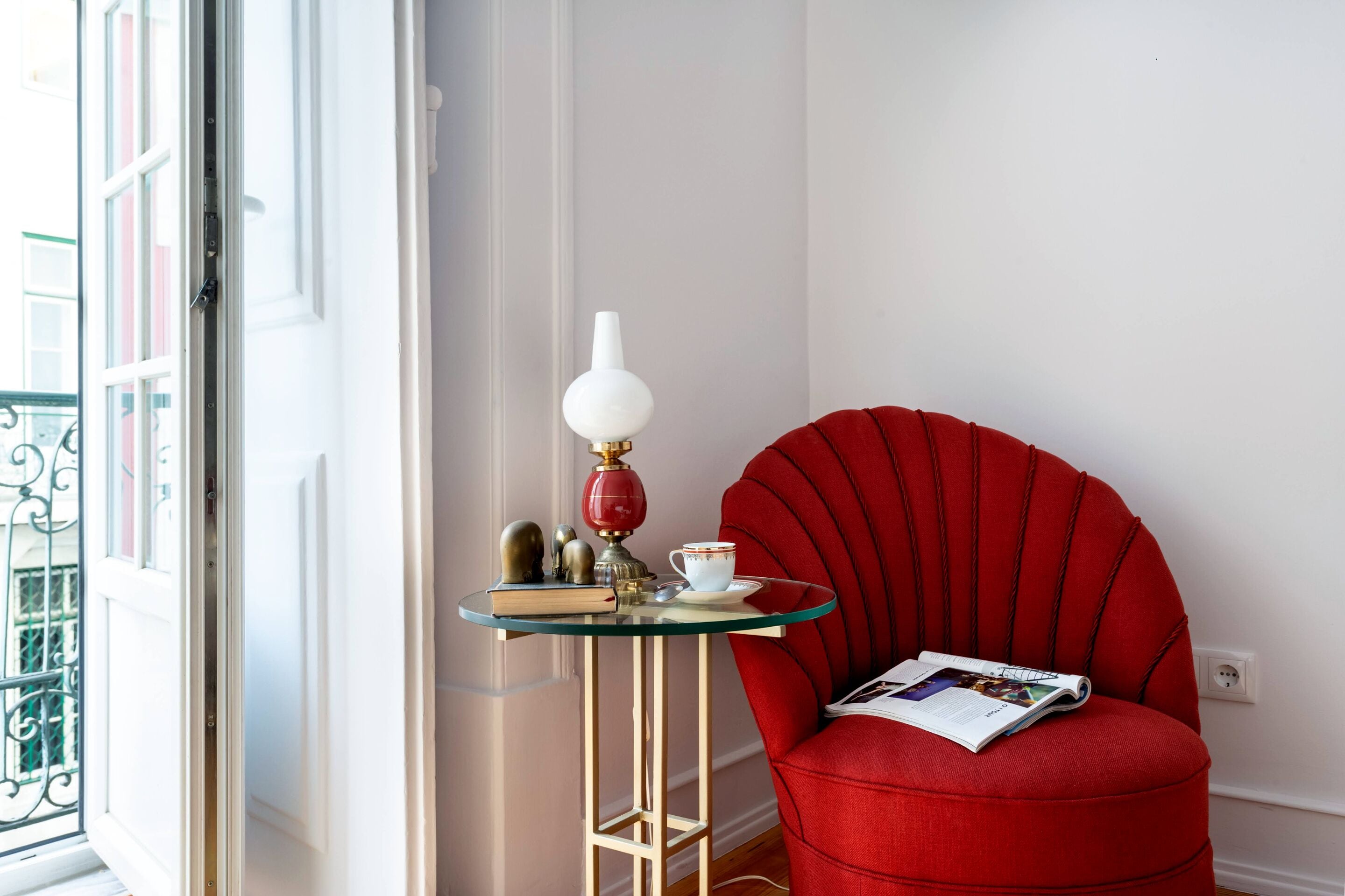 A red armchair with a magazine, a glass table with a lamp, and a window with a view of buildings.