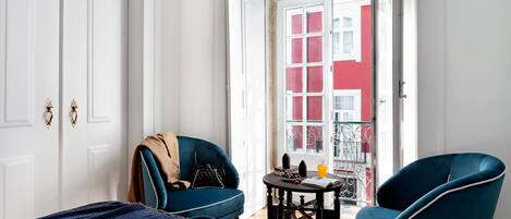 A room with a bed, two blue armchairs, a small table with a book and a glass of orange juice, and a view of a red building through a glass door.