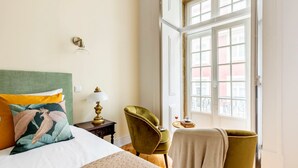 A hotel room with a bed, a green armchair, a lamp, and a window with a view of buildings. - LeL Madalena GuestHouse (Lisbon)