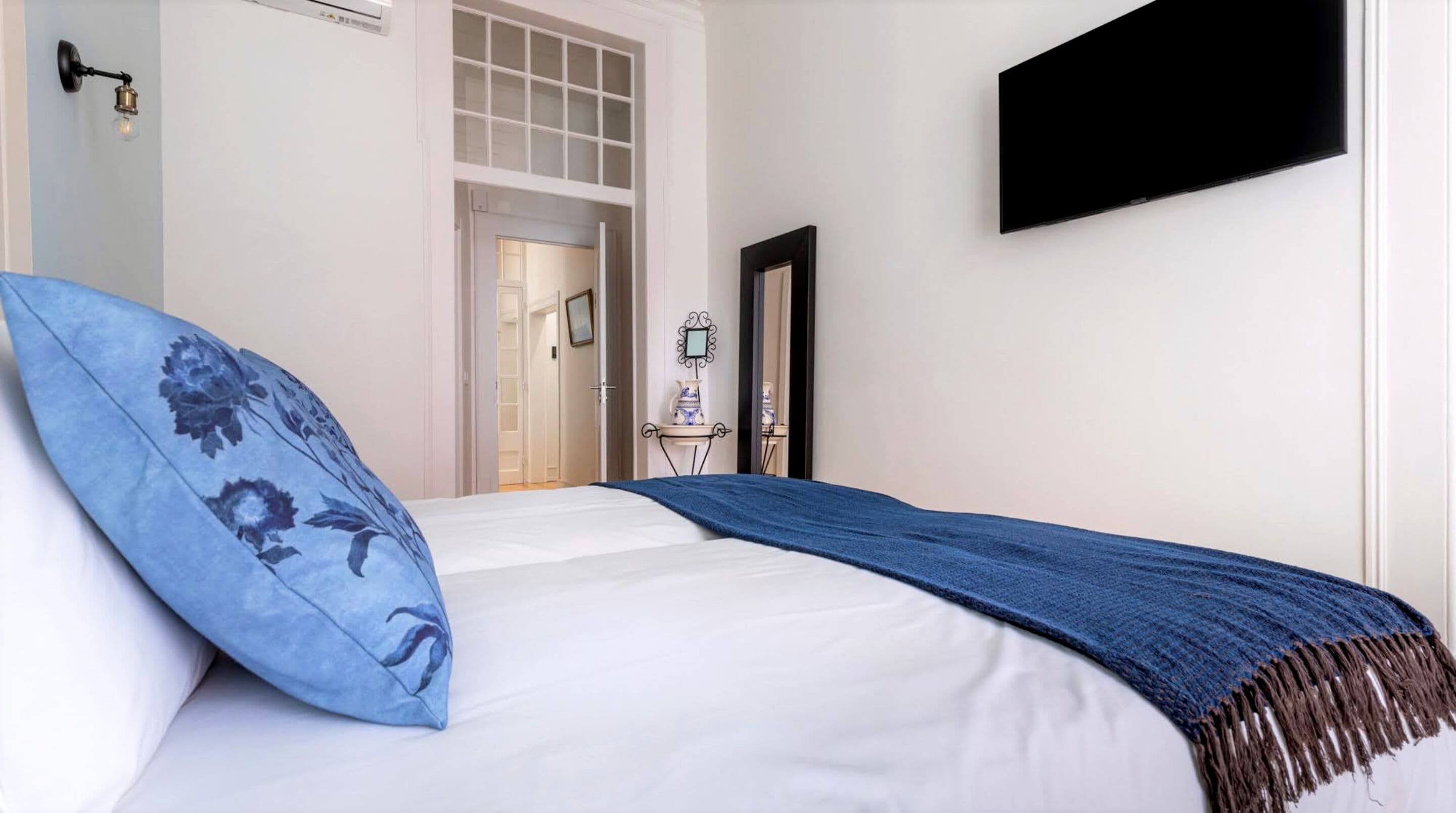 Un dormitorio con cama, un cojín floral azul, una manta azul, un televisor de pantalla plana montado en la pared y una mesita de noche con adornos.
