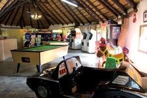 Uma mesa de bilhar, máquinas de fliperama e uma máquina caça-níqueis em formato de carro antigo em um ambiente com teto alto e vigas de madeira.