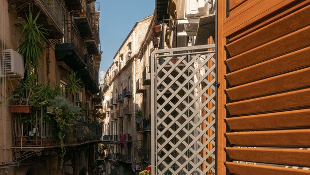 A narrow street with buildings on either side, a wooden gate, and a planter with flowers. - San Francesco Rooms L.T. (Palermo)