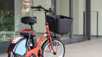A red electric bike with a black basket and a rear storage box, parked on a sidewalk.