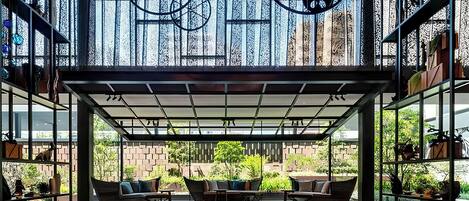A modern outdoor seating area with a glass enclosure, furnished with sofas and tables, overlooking a brick wall and greenery.