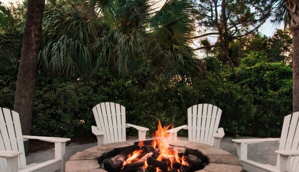 A fire pit with white chairs around it, set in a garden with dense foliage and trees.