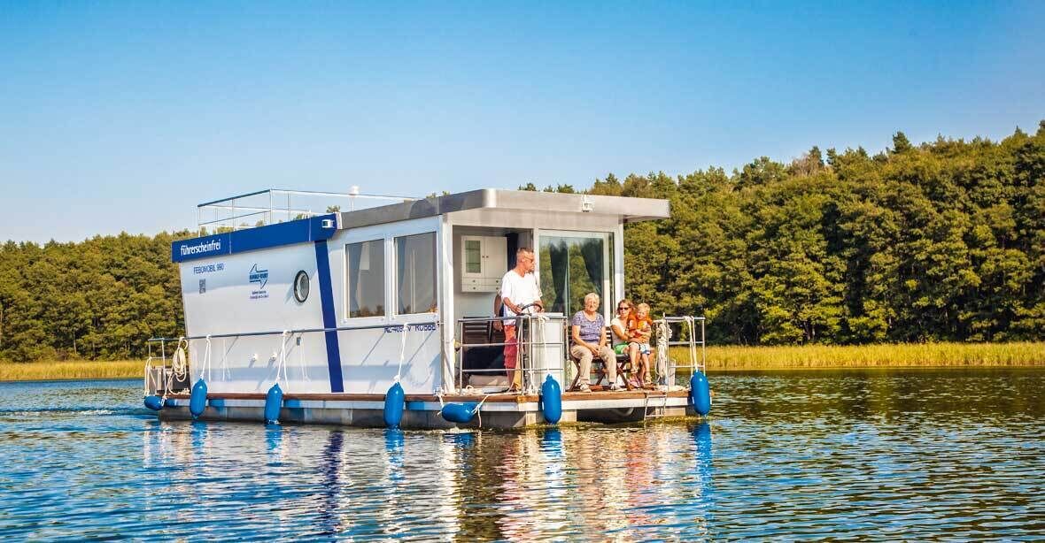 A houseboat with a covered deck and a small cabin, floating on a calm lake with a forested shoreline in the background.