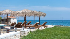 A beachside lounge area with wooden loungers and thatched umbrellas, a stone wall, and a view of the sea. - Musela Studios (Rethymno)