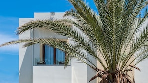 A modern two-story building with a balcony, a palm tree, and a view of the sea. - Musela Studios (Rethymno)